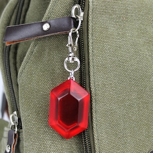 May include: A red, octagonal-shaped keychain charm attached to a silver key ring. The charm is hanging from a brown leather strap on a green canvas backpack.