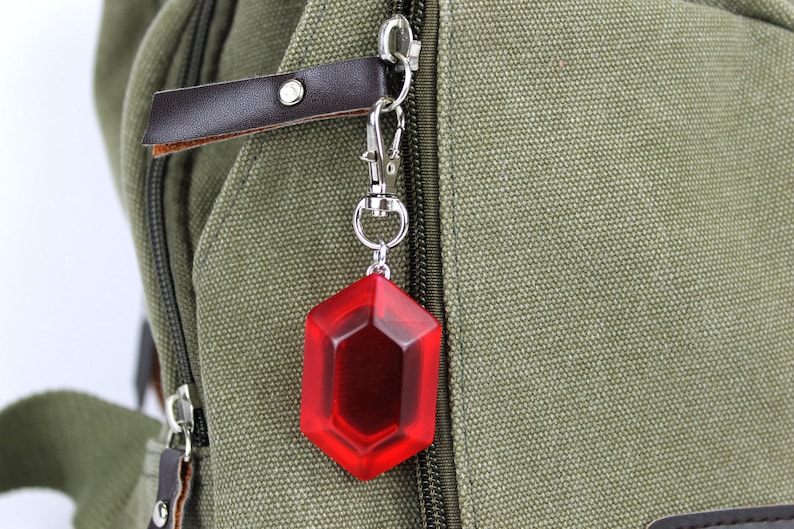 May include: A red, octagonal-shaped keychain charm attached to a silver key ring. The charm is hanging from a brown leather strap on a green canvas backpack.