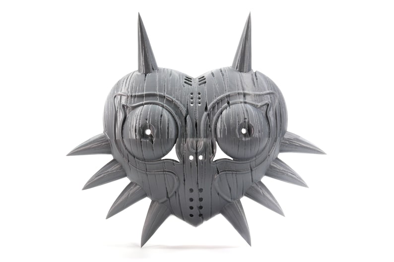 May include: A gray, heart-shaped mask with sharp, pointed spikes around the edges. The mask has two large, round eye holes and a smaller hole in the center. The mask is made of a textured material that resembles wood.