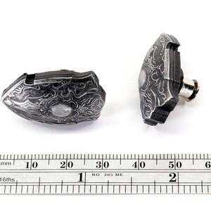 May include: Two silver-coloured metal shield-shaped buttons with intricate designs. The buttons are approximately 2 cm in diameter and have a pin on the back for attaching to fabric.