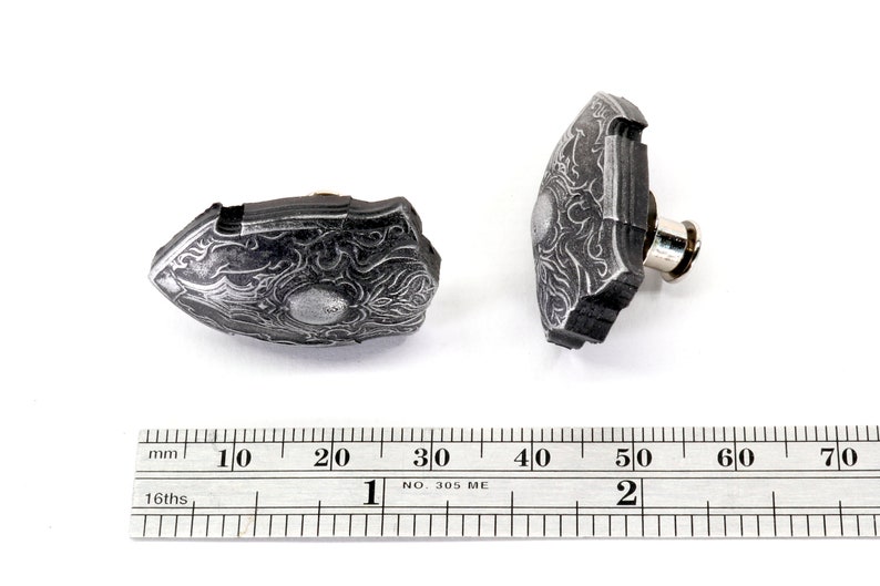 May include: Two silver-coloured metal shield-shaped buttons with intricate designs. The buttons are approximately 2 cm in diameter and have a pin on the back for attaching to fabric.