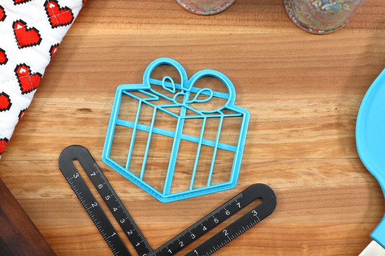 Op de afbeelding: Een lichtblauwe plastic koekjesvormer in de vorm van een cadeaudoos met een strik erop. De koekjesvormer ligt op een houten oppervlak.