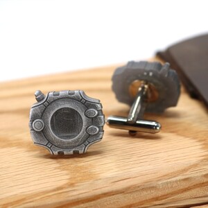 May include: A pair of silver cufflinks with a detailed design. The cufflinks are on a wooden surface.