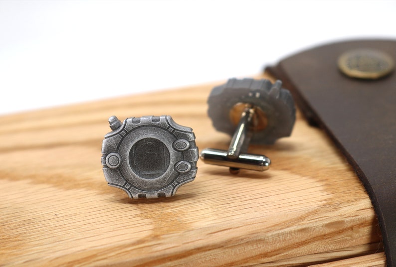 May include: A pair of silver cufflinks with a detailed design. The cufflinks are on a wooden surface.