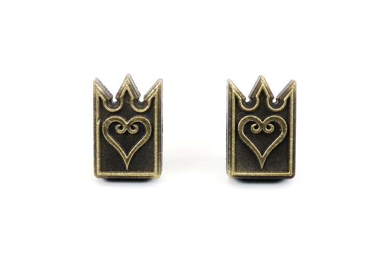 May include: Two bronze-colored rectangular earrings with a crown design at the top and a heart in the center. The earrings are set against a white background.