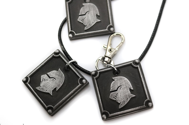 May include: Three silver-toned metal pendants with a knight's helmet design. The pendants are attached to a black cord and a silver-toned metal clasp.