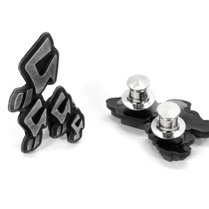 May include: Two black metal pins with a silver metal clasp. The pins are shaped like a stylized letter 'G' and have a textured surface.