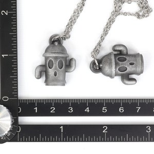 May include: Two silver necklaces with robot-like pendants. The pendants are shaped like a robot with a round head, two arms, and two legs. The necklaces are on a white background.