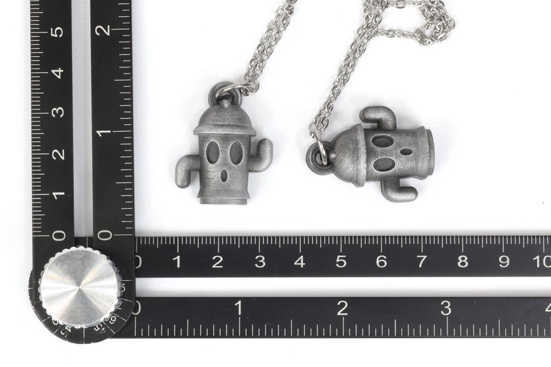 May include: Two silver necklaces with robot-like pendants. The pendants are shaped like a robot with a round head, two arms, and two legs. The necklaces are on a white background.