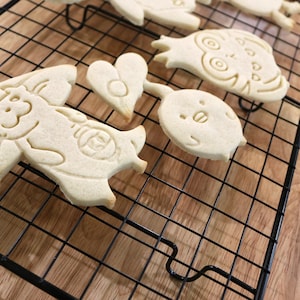 May include: Five sugar cookies on a black wire cooling rack. The cookies are in the shapes of a heart, a penguin, an owl, a cat, and a cat wearing a hat. The cookies are on a wooden surface.
