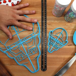 May include: Two blue plastic cookie cutters shaped like a cartoon face with a hat. The larger cutter is being held by a hand. The smaller cutter is on a wooden surface. There is a ruler and a spatula in the background.