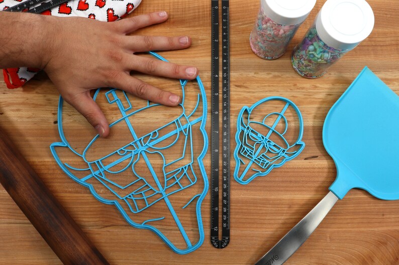 May include: Two blue plastic cookie cutters shaped like a cartoon face with a hat. The larger cutter is being held by a hand. The smaller cutter is on a wooden surface. There is a ruler and a spatula in the background.