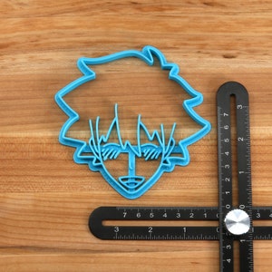 May include: A blue plastic cookie cutter shaped like a cartoon character with spiky hair and sunglasses. The cutter is approximately 2 inches wide.