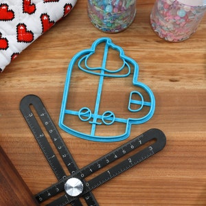 May include: A blue plastic cookie cutter shaped like a mug with a swirl of cream on top. The cutter is on a wooden surface with a ruler and a rolling pin.