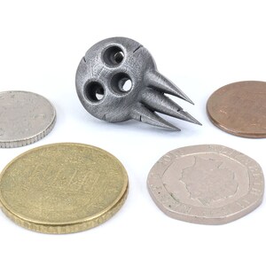 Op de afbeelding: Een zilverkleurig metalen schedelvormig object met drie gaten en scherpe punten. Het object wordt omringd door vier munten.