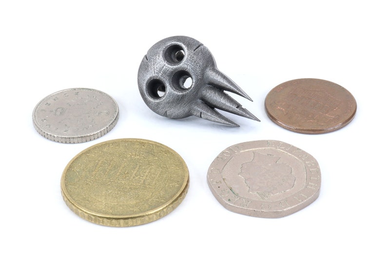 Op de afbeelding: Een zilverkleurig metalen schedelvormig object met drie gaten en scherpe punten. Het object wordt omringd door vier munten.