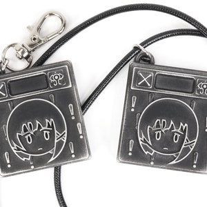May include: Two silver-colored keychains with black cords. Each keychain features a square metal charm with a cartoon face and exclamation points around the border. The charms also have a black button and a symbol that looks like an eye.