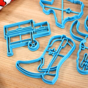 May include: A set of blue plastic cookie cutters shaped like a cowboy boot, a grill, a longhorn skull, a cowboy hat, and the state of Texas.