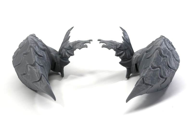 Op de afbeelding: Een paar grijze, 3D-geprinte vleugels met een textuur. De vleugels zijn symmetrisch en hebben een puntige vorm.