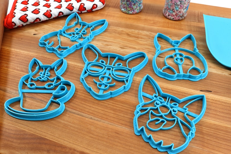 Pu&ograve; includere: Cinque tagliabiscotti in plastica blu a forma di corgi. I tagliabiscotti presentano diverse espressioni, tra cui uno con gli occhiali, uno che tiene una tazza di caff&egrave; e uno con la lingua di fuori.