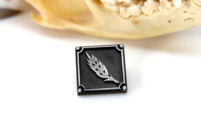 May include: A silver wheat stalk design on a black square metal pin.
