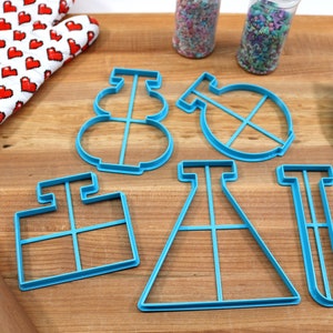 May include: A set of five blue plastic cookie cutters shaped like a snowman, a beaker, a test tube, a flask, and a square. The cutters are on a wooden surface with a rolling pin, a spatula, and a red oven mitt.