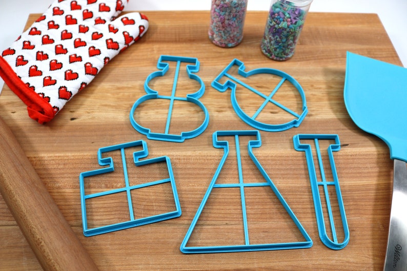 May include: A set of five blue plastic cookie cutters shaped like a snowman, a beaker, a test tube, a flask, and a square. The cutters are on a wooden surface with a rolling pin, a spatula, and a red oven mitt.
