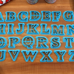 May include: A set of 26 blue plastic alphabet cookie cutters on a wooden surface. The cutters are in uppercase letters and are arranged in alphabetical order.