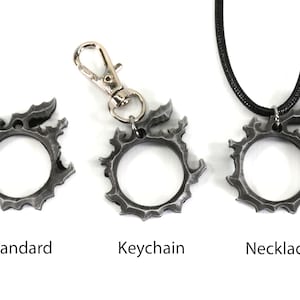 Puede incluir: Tres versiones de un colgante de metal de color plateado con forma de c&iacute;rculo con bordes dentados. El colgante se muestra como una pieza est&aacute;ndar, un llavero y un collar.