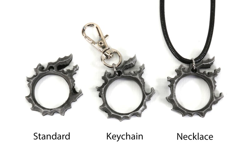 Puede incluir: Tres versiones de un colgante de metal de color plateado con forma de c&iacute;rculo con bordes dentados. El colgante se muestra como una pieza est&aacute;ndar, un llavero y un collar.