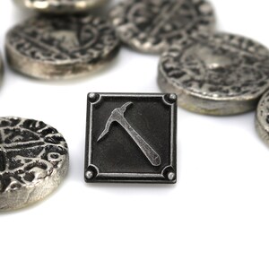 K&ouml;nnte beinhalten: Eine schwarze Metallnadel mit einem silbernen Pickel-Design. Die Nadel ist von mehreren silbernen M&uuml;nzen mit komplizierten Designs umgeben.
