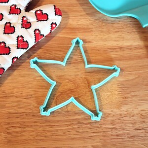 Pu&ograve; includere: Un tagliabiscotti a forma di stella azzurra su una superficie di legno, accanto a una spatola blu abbinata e a un guanto da forno a forma di cuore. Il guanto da forno ha uno sfondo bianco con cuori pixelati rossi.