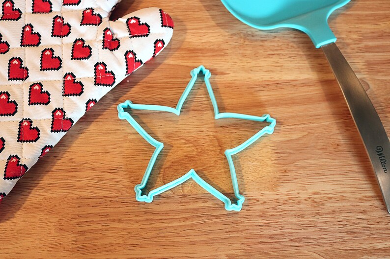 Pu&ograve; includere: Un tagliabiscotti a forma di stella azzurra su una superficie di legno, accanto a una spatola blu abbinata e a un guanto da forno a forma di cuore. Il guanto da forno ha uno sfondo bianco con cuori pixelati rossi.