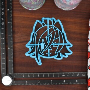 May include: A blue plastic cookie cutter shaped like a cartoon character with a large eye and long hair. The cutter is on a wooden surface.