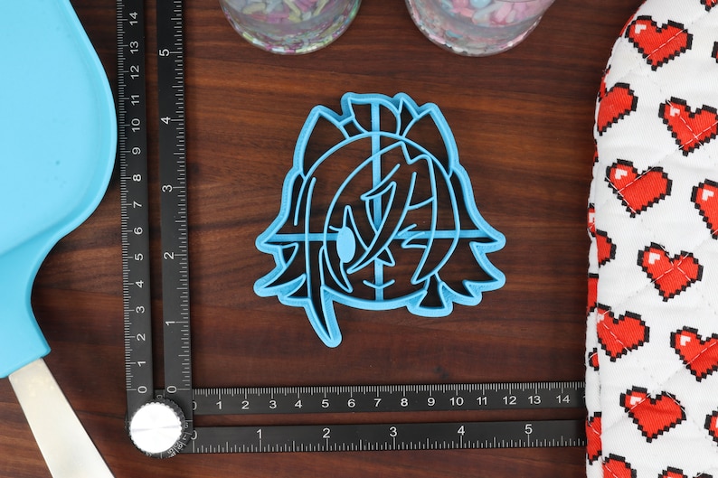 May include: A blue plastic cookie cutter shaped like a cartoon character with a large eye and long hair. The cutter is on a wooden surface.