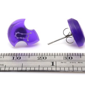 May include: A pair of purple stud earrings with a Gemini zodiac symbol on one earring. The earrings are shown next to a ruler for size reference.