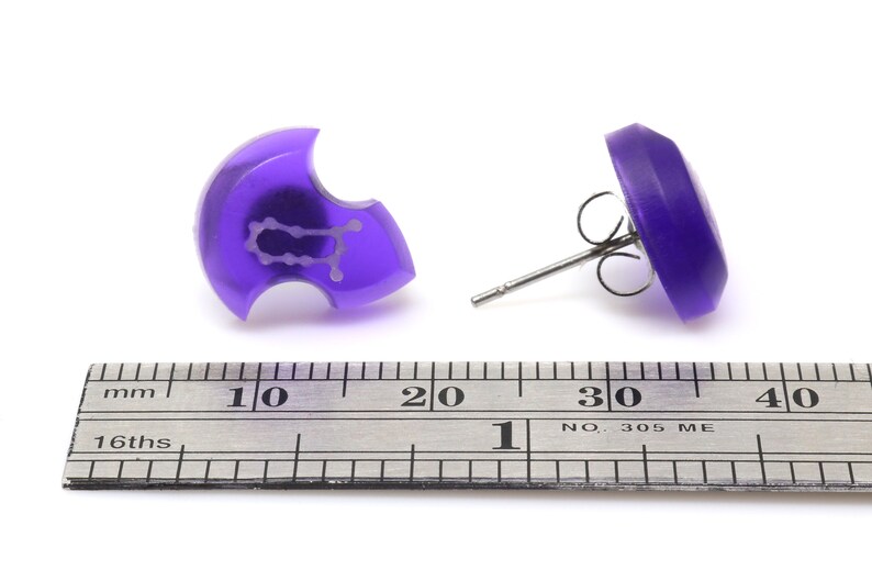 May include: A pair of purple stud earrings with a Gemini zodiac symbol on one earring. The earrings are shown next to a ruler for size reference.