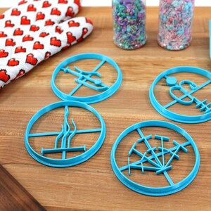 May include: Four blue plastic cookie cutters shaped like different designs on a wooden surface. The cutters are for making cookies in the shape of a tree, a lighthouse, a palm tree, and a compass.