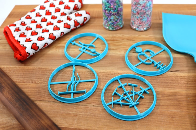 May include: Four blue plastic cookie cutters shaped like different designs on a wooden surface. The cutters are for making cookies in the shape of a tree, a lighthouse, a palm tree, and a compass.