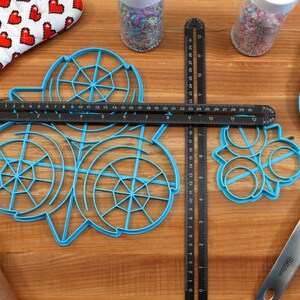 May include: A blue plastic cookie cutter set with three different designs. The set includes a circle with a web design, a circle with a swirl design, and a circle with a flower design. The cookie cutters are on a wooden surface with a ruler, a rolling pin, and a spatula.