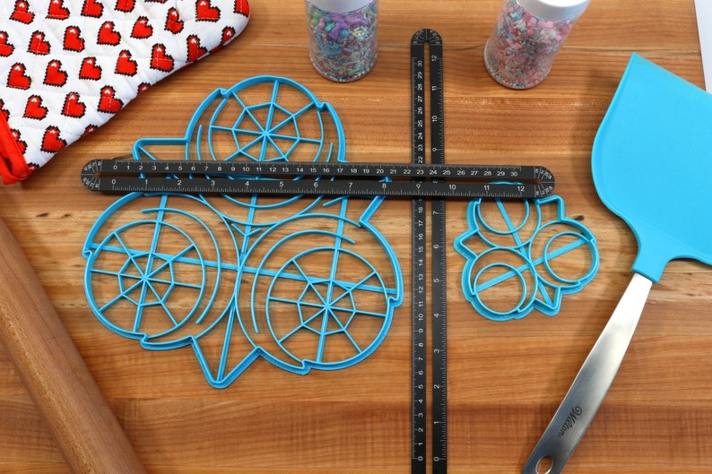 May include: A blue plastic cookie cutter set with three different designs. The set includes a circle with a web design, a circle with a swirl design, and a circle with a flower design. The cookie cutters are on a wooden surface with a ruler, a rolling pin, and a spatula.