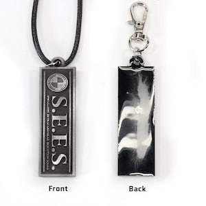 May include: A silver-colored necklace with a rectangular pendant that reads "S.E.E.S. SPECIAL EXTRATERRESTRIAL ENFORCEMENT SQUAD" with a circle containing a cross inside. The back of the pendant is blank and silver.