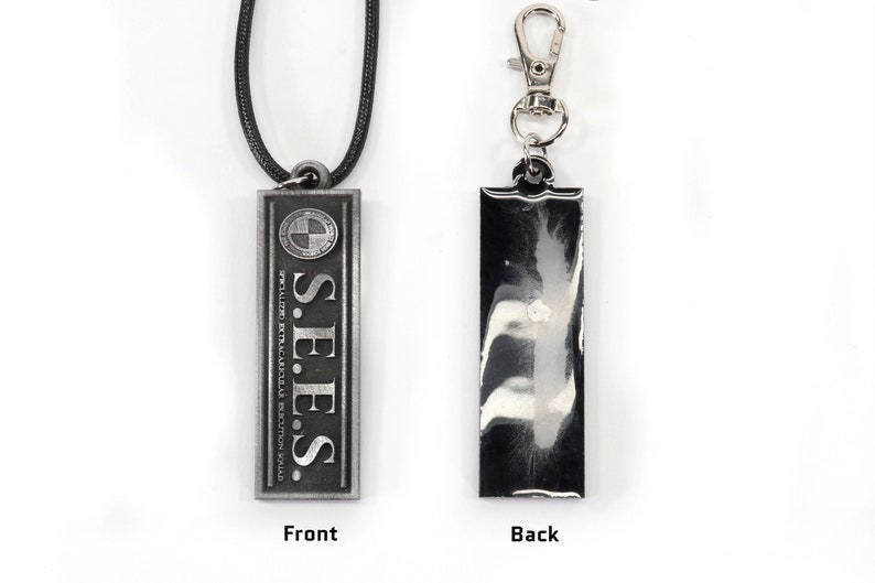 May include: A silver-colored necklace with a rectangular pendant that reads "S.E.E.S. SPECIAL EXTRATERRESTRIAL ENFORCEMENT SQUAD" with a circle containing a cross inside. The back of the pendant is blank and silver.