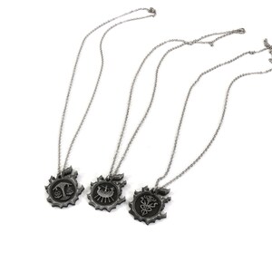 May include: Three silver necklaces with different pendants. Each pendant features a stylized design with a black finish. The pendants are shaped like a circle with a gear-like border.