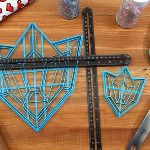 Puede incluir: Dos cortadores de galletas de pl&aacute;stico azul con forma de cristales geom&eacute;tricos. El cortador m&aacute;s grande mide 76 cm de largo y el m&aacute;s peque&ntilde;o 30 cm. Los cortadores est&aacute;n sobre una superficie de madera con una regla, un rodillo, una esp&aacute;tula y un frasco de chispas.