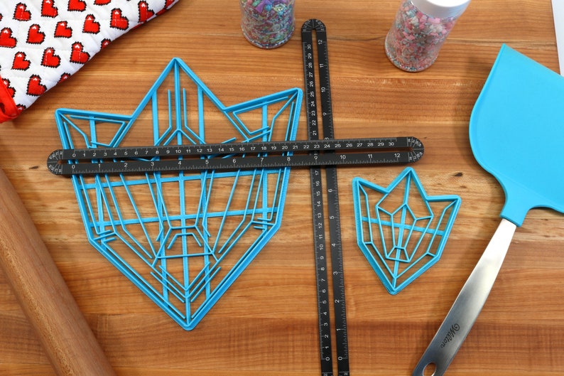Puede incluir: Dos cortadores de galletas de pl&aacute;stico azul con forma de cristales geom&eacute;tricos. El cortador m&aacute;s grande mide 76 cm de largo y el m&aacute;s peque&ntilde;o 30 cm. Los cortadores est&aacute;n sobre una superficie de madera con una regla, un rodillo, una esp&aacute;tula y un frasco de chispas.