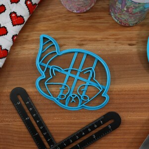 Puede incluir: Cortador de galletas de pl&aacute;stico azul con forma de mapache con un gorro de fiesta. El cortador tiene un patr&oacute;n de cuadr&iacute;cula en el interior.
