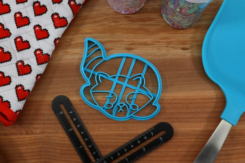 Puede incluir: Cortador de galletas de pl&aacute;stico azul con forma de mapache con un gorro de fiesta. El cortador tiene un patr&oacute;n de cuadr&iacute;cula en el interior.