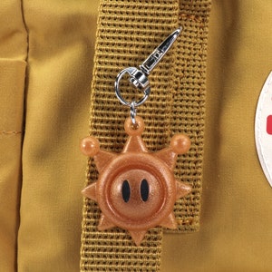 May include: A gold-colored sun charm with black eyes attached to a silver key chain. The charm is hanging from a brown backpack strap.