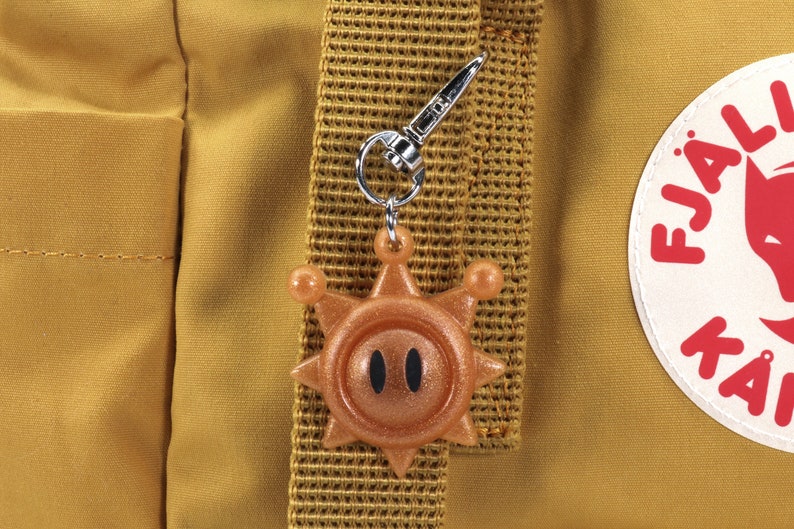 May include: A gold-colored sun charm with black eyes attached to a silver key chain. The charm is hanging from a brown backpack strap.